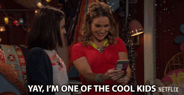 two women are looking at a cell phone and one of them says yay i 'm one of the cool kids