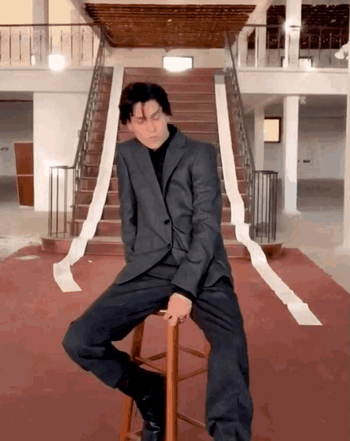 a man in a suit sits on a wooden stool in front of a set of stairs