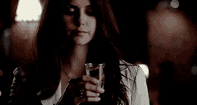 a woman in a white jacket is drinking from a glass in a dark room .