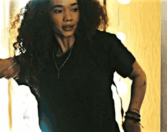 a woman with curly hair wearing a black t-shirt and a necklace is standing in front of a mirror .