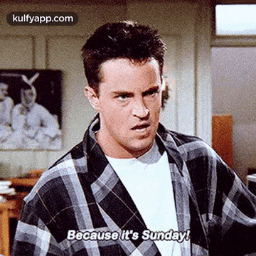 a man in a plaid shirt is standing in a room and says `` because it 's sunday ! ''