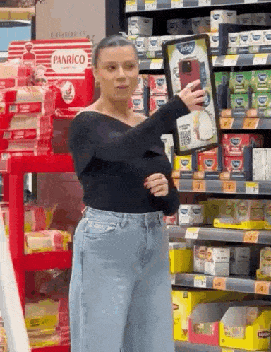 a woman in a black top and blue jeans is standing in a grocery store holding a sign .