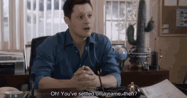 a man in a blue shirt is sitting at a desk and says oh you 've settled on a name then?