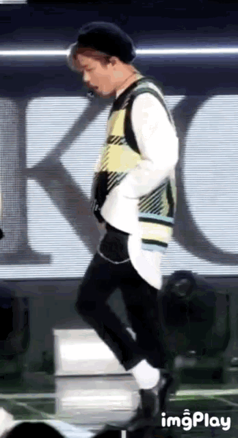 a man in a plaid vest is walking on a stage with the word imgplay behind him