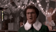 a man in a green shirt and white collar is standing in front of a christmas tree with lights .