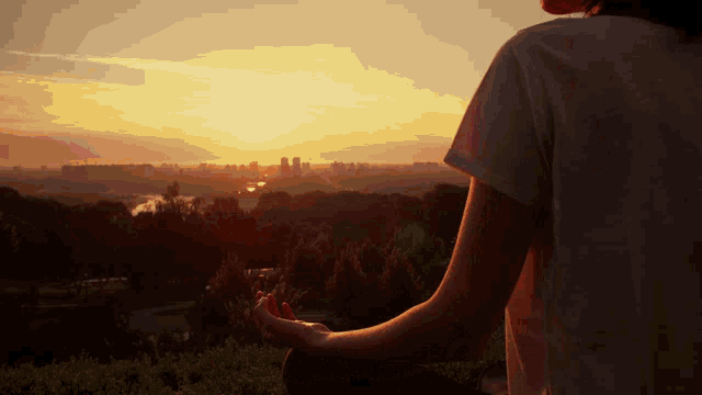 a woman sits in a lotus position looking at the sunset over a city
