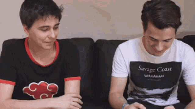 two men are sitting on a couch and one has a savage coast shirt on