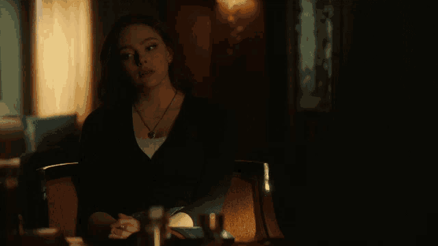 a woman sits at a table in a dark room wearing a heart shaped necklace