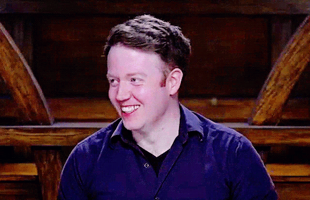 a man in a blue shirt is smiling in front of a wooden ceiling