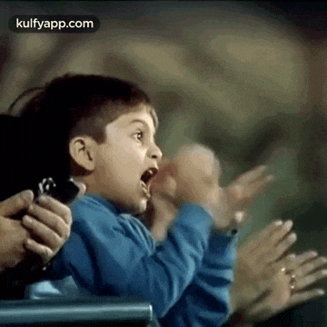a young boy is holding a cell phone in his hand while sitting in a crowd of people .