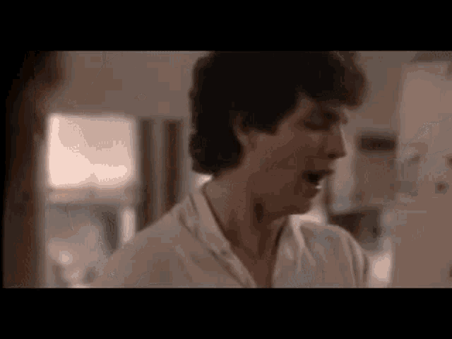 a man in a white shirt is standing in a kitchen talking to someone .