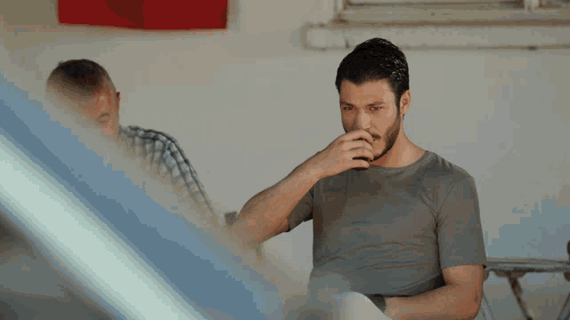 a man covering his nose while another man sits behind him
