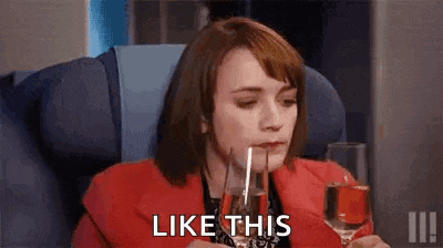 a woman is sitting on an airplane holding a glass of wine and says `` like this '' .