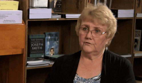 a woman is sitting in front of a bookshelf with a book titled the gospel according to joseph smith