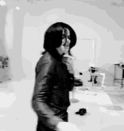a black and white photo of a man in a leather jacket standing in a room talking on a cell phone .