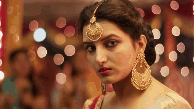 a woman wearing gold earrings and a maang tikka looks at the camera