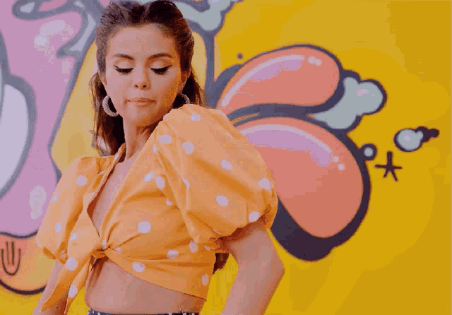 a woman wearing a polka dot top and hoop earrings stands in front of a yellow wall