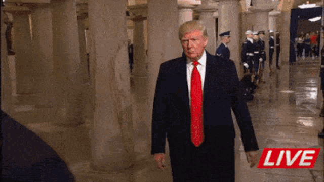 a man in a suit and red tie is standing in a hallway with soldiers behind him and a live sign in the corner