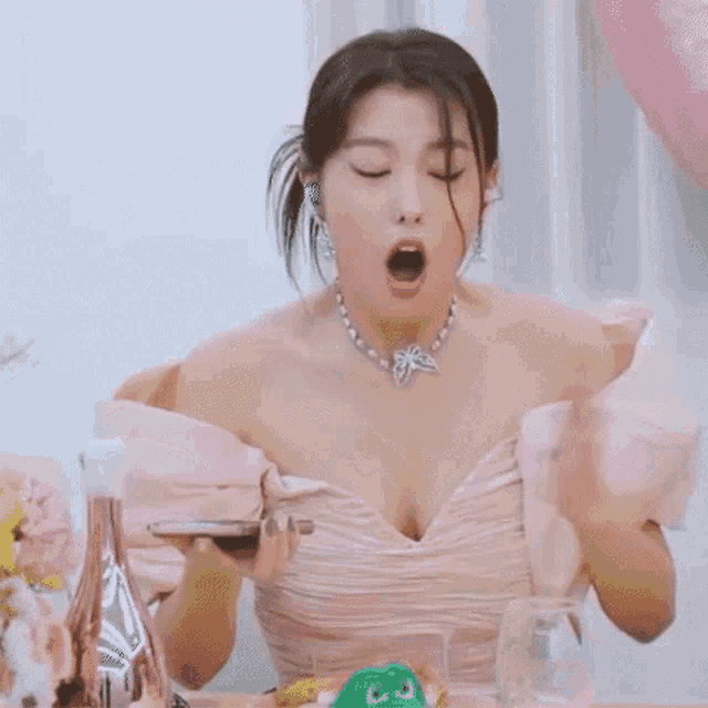 a woman in a pink dress and necklace is sitting at a table with her hands on her chest .