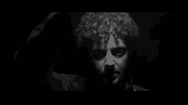a black and white photo of a man with curly hair and a beard in a dark room .