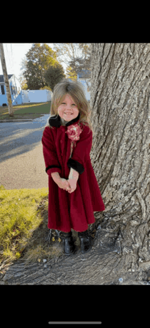 a little girl wearing a red coat and black boots stands next to a tree