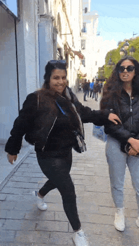 two women are walking down a sidewalk and one of them is wearing sunglasses .