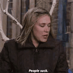 a woman in a black jacket is sitting in front of a tree and saying `` people suck '' .
