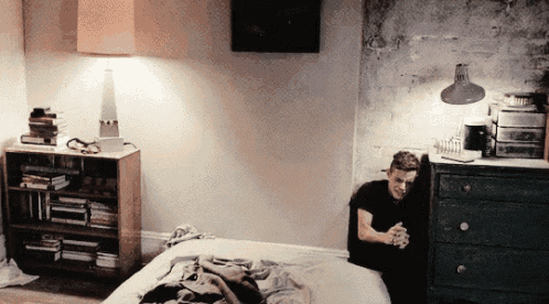 a man is sitting on a bed in a bedroom with a lamp and a dresser