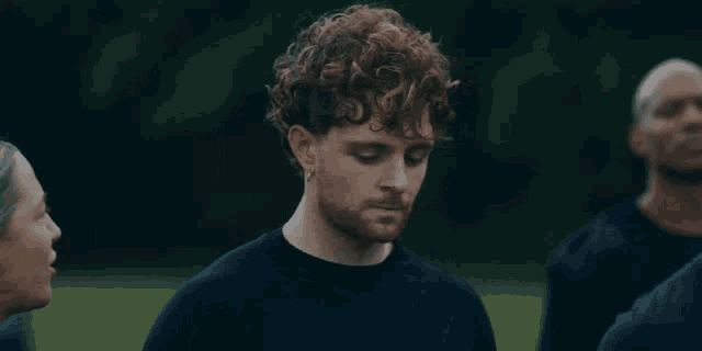 a man with curly hair and a tattoo on his chest talks to a woman