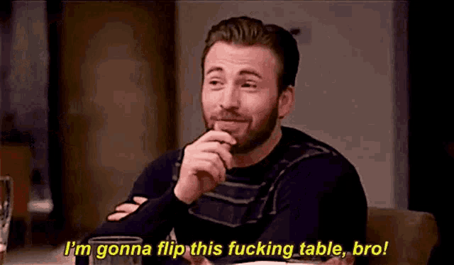 a man with a beard is sitting at a table with a glass of beer and talking .