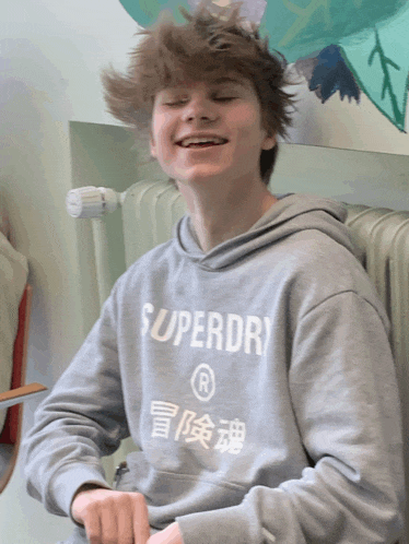 a young man wearing a grey superdry sweatshirt smiles with his eyes closed