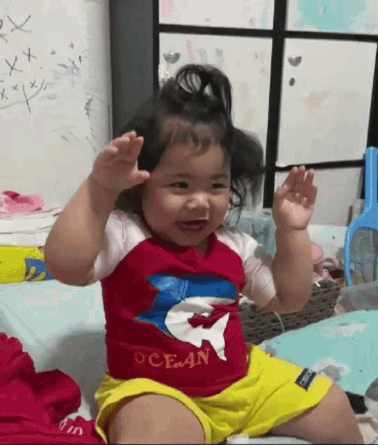 a baby girl wearing a red shirt with a whale on it is sitting on a bed .