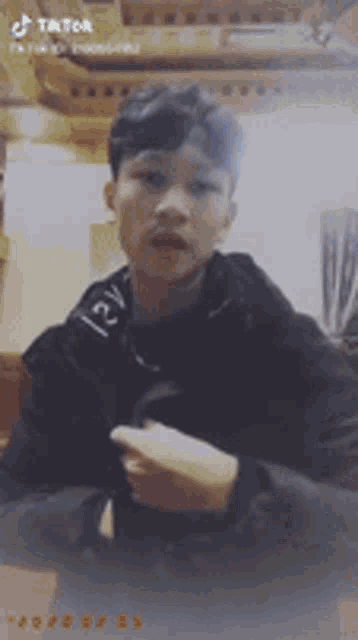 a young man in a black jacket is sitting at a table and making a funny face .