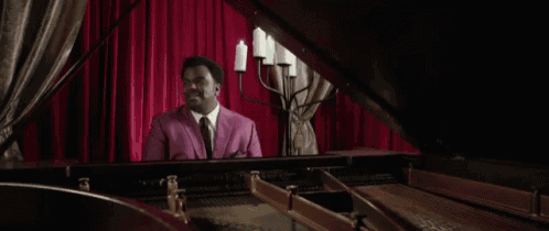 a man in a pink suit playing a piano in front of a red curtain