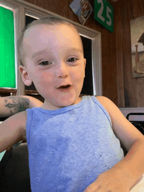 a little boy in a blue tank top is sitting in front of a green sign with the number 25 on it