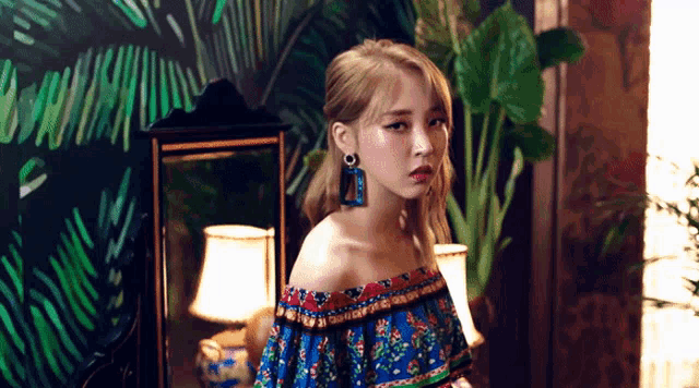 a woman wearing a blue off the shoulder top is standing in front of a mirror and a lamp .