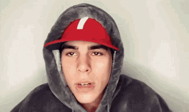 a young man wearing a hooded sweatshirt and a red hat with the letter t on it .