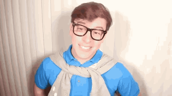 a young man wearing glasses , a blue shirt , and a gray sweater is smiling .