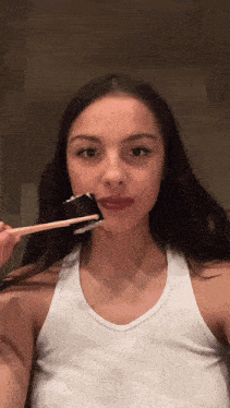a woman in a white tank top is brushing her teeth