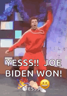 a man in a red shirt is dancing with his arms in the air and says yesss ! joe biden won !