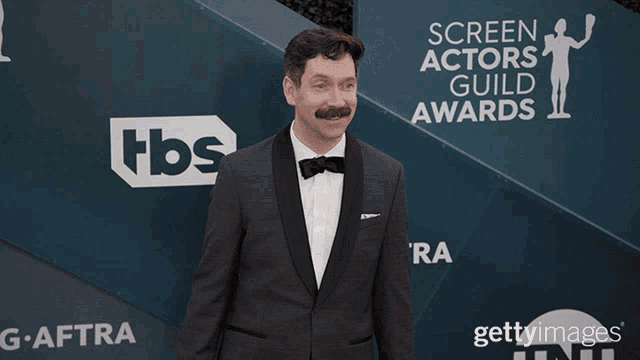 a man in a tuxedo stands in front of a screen actors guild awards banner
