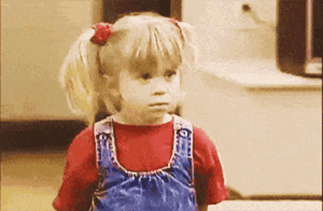 a little girl wearing overalls and a red shirt is making a face
