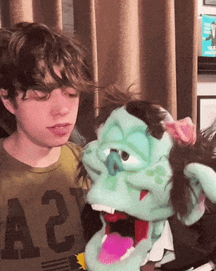 a boy wearing a green shirt with the letter a on it looks at a puppet