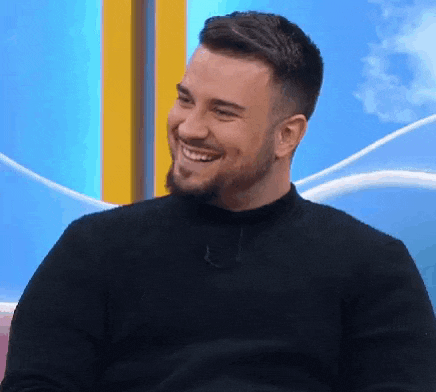 a man with a beard wearing a black sweater is smiling while sitting on a chair .