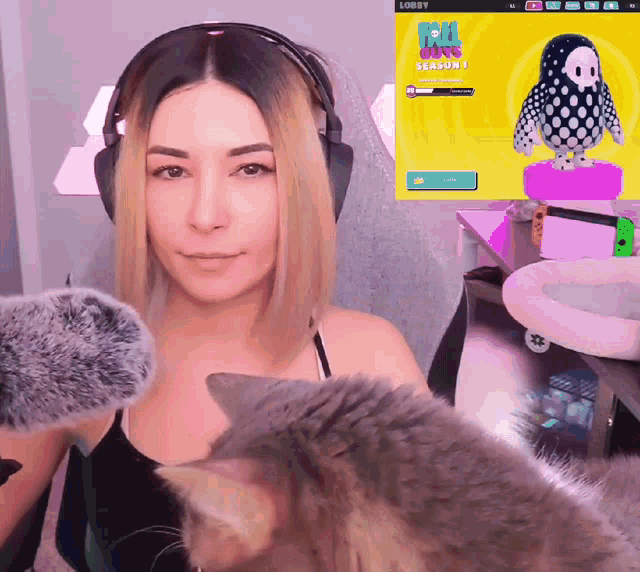 a woman wearing headphones looks at a cat in front of a screen that says fall guys