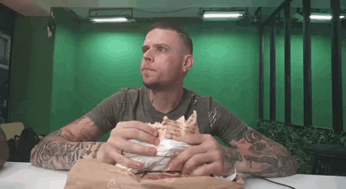 a man with tattoos is sitting at a table eating a large sandwich .