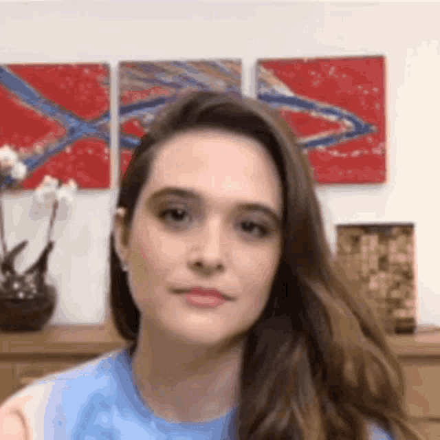 a woman in a blue shirt is looking at the camera in front of a wall with paintings .