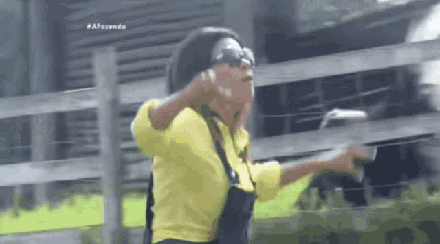 a woman in a yellow shirt and black overalls is standing in front of a fence .