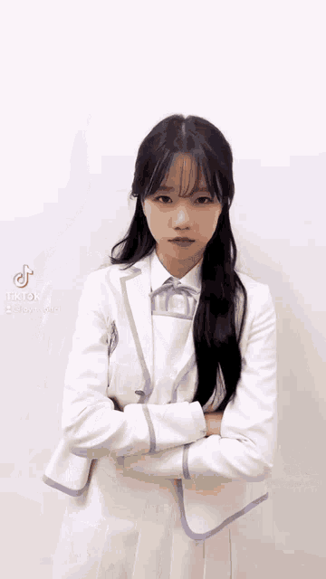 a girl in a white suit and purple collared shirt is standing with her arms crossed .