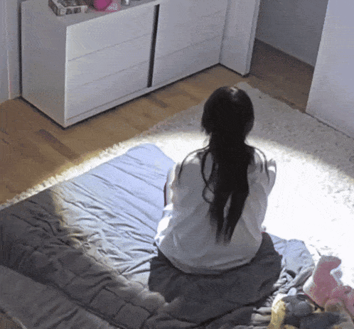 a woman sits on a bed with her back to the camera and a book on the dresser behind her